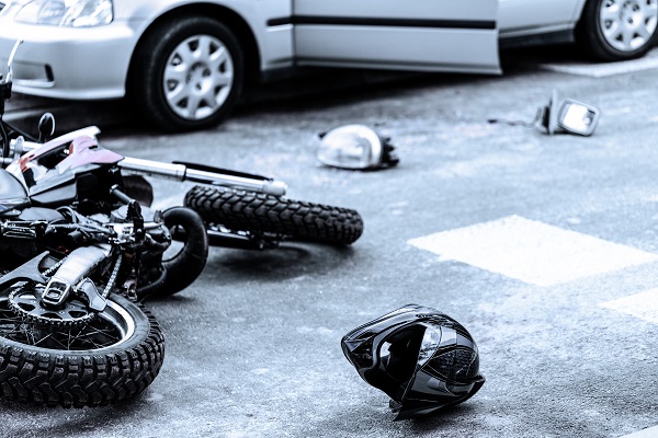 Damaged motorcycle lying on the pavement next to a car with its door open, surrounded by debris including a helmet and broken mirror, suggesting a serious crash.