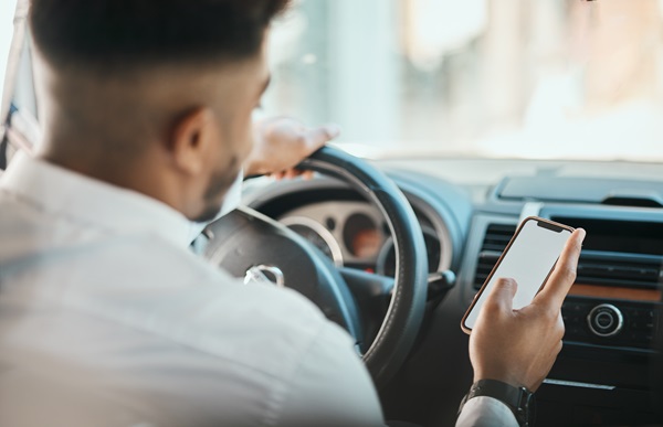 Man driving a car while looking down at his smartphone, with one hand on the wheel and the other holding the device.