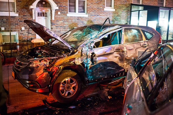 Severely damaged SUV at night with a crumpled hood, shattered windows, and a dented side, sitting on a city street near a brick apartment building.